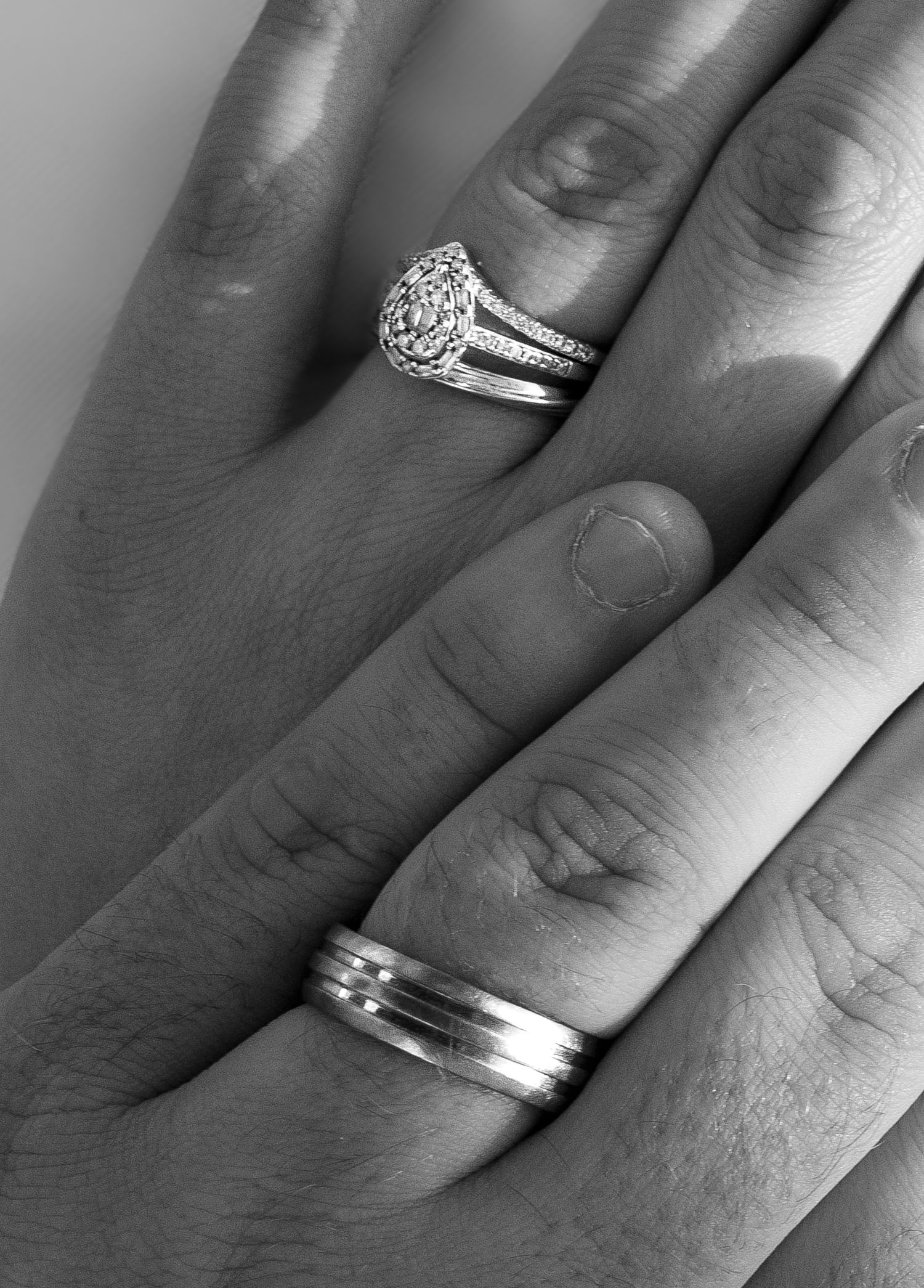 Black and white photo of two hands displaying wedding rings, one with a diamond halo engagement ring and one with a mens band.