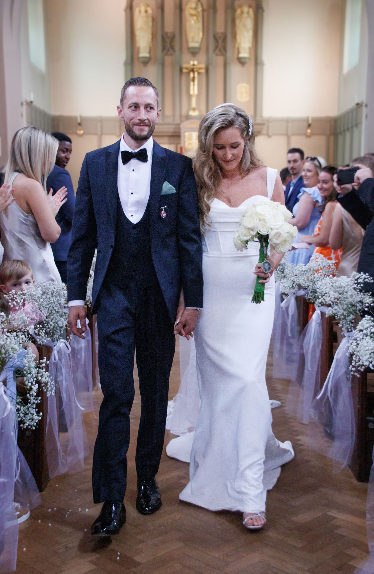Wedding couple walks down church aisle decorated with white flowers and ribbons.