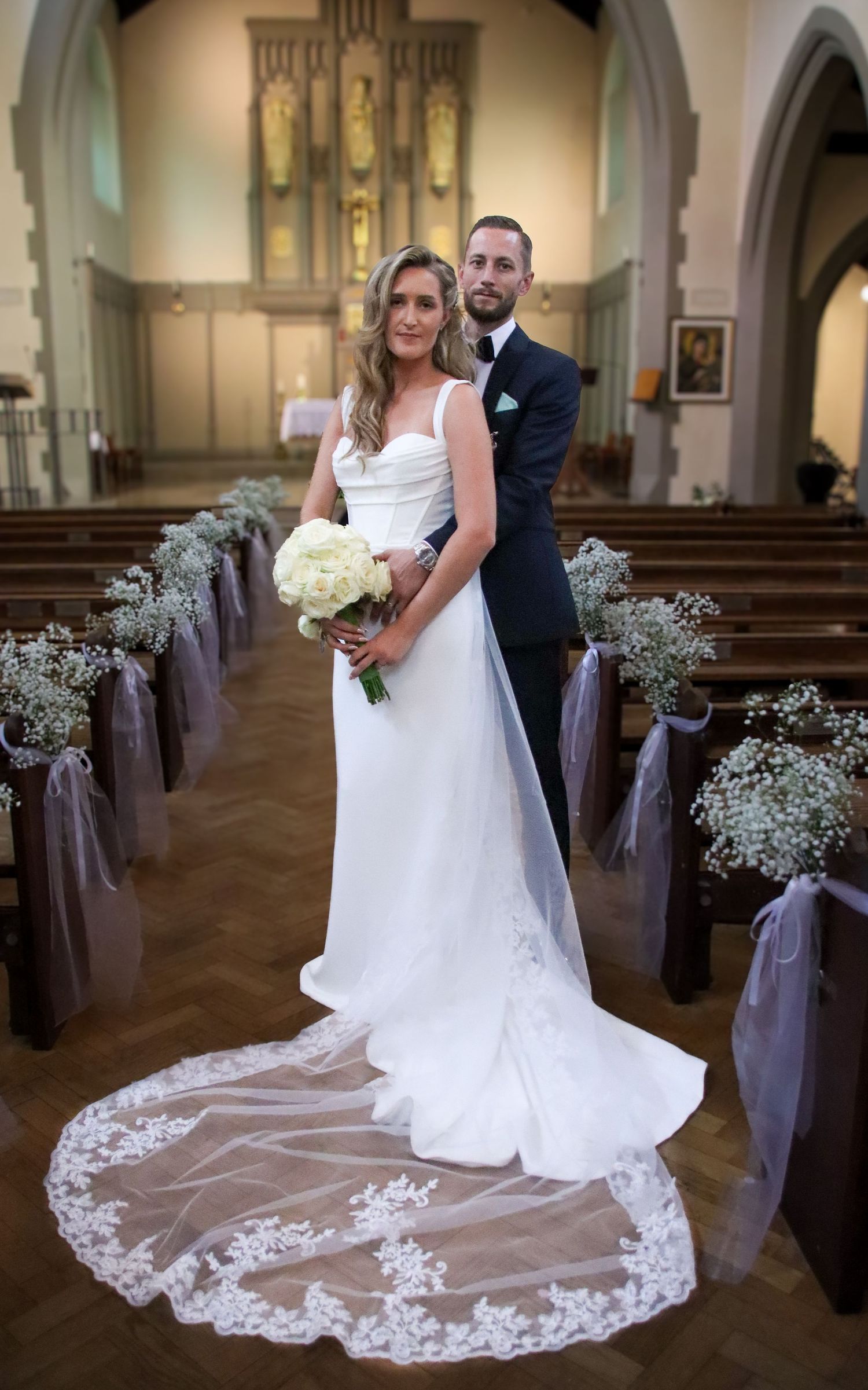Wedding couple poses in a traditional church with decorated pews and a long lace-trimmed veil.