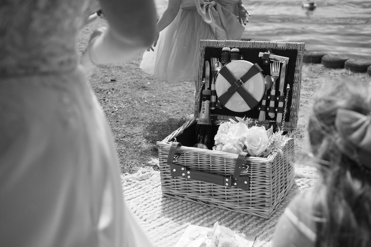 Black and white photo of an open wicker picnic basket filled with bottles and flowers on a blanket outdoors.