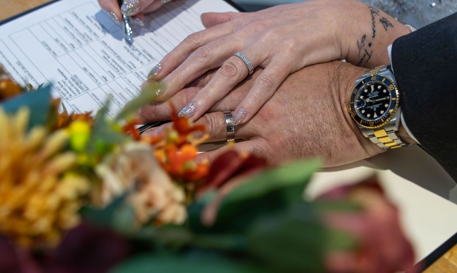 Close-up of hands with gold watch and rings signing documents next to colorful food platter.