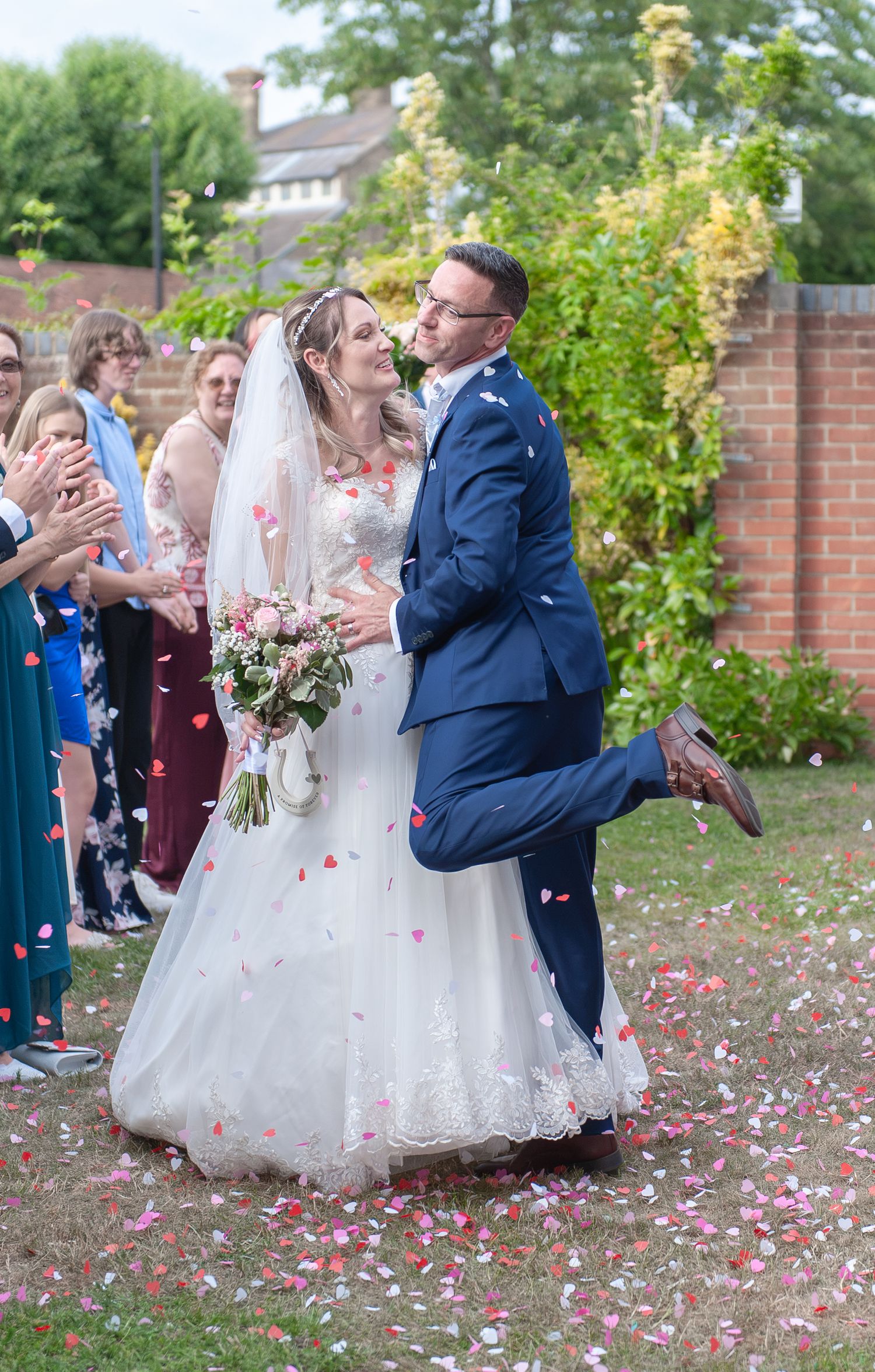 Bride and groom share a kiss during confetti shower at outdoor wedding ceremony, guests applauding in background.