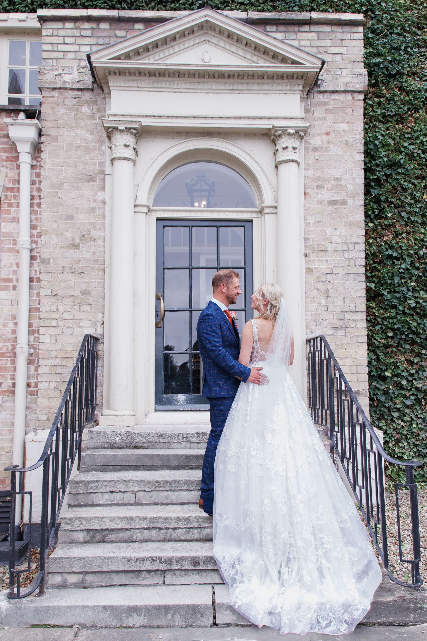 Bride and groom stand on stone steps before an elegant arched doorway, her lace gown trailing beautifully.
