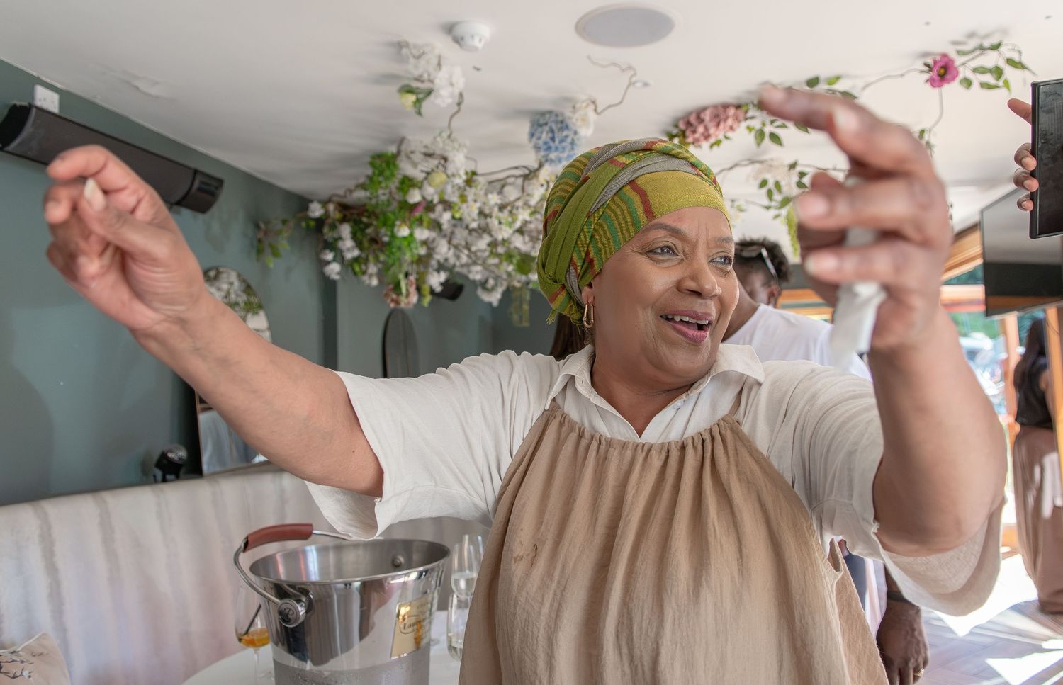 A joyful woman in a green headwrap gestures expressively while cooking in a floral-decorated kitchen.
