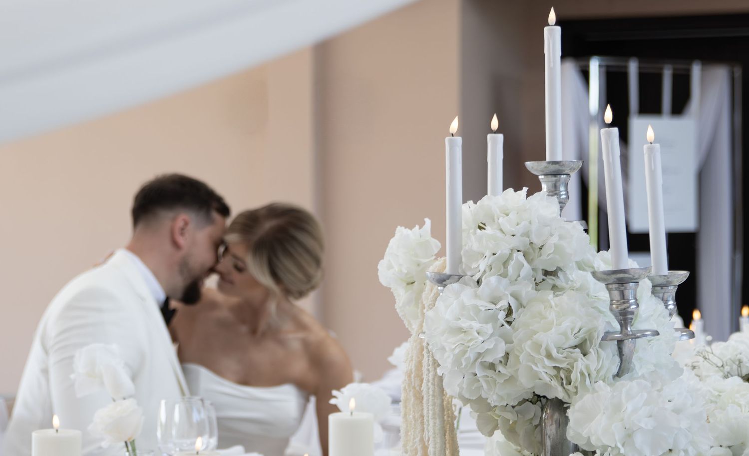 Bride and groom sharing a kiss at elegant wedding reception with white floral centerpiece and lit candelabra.