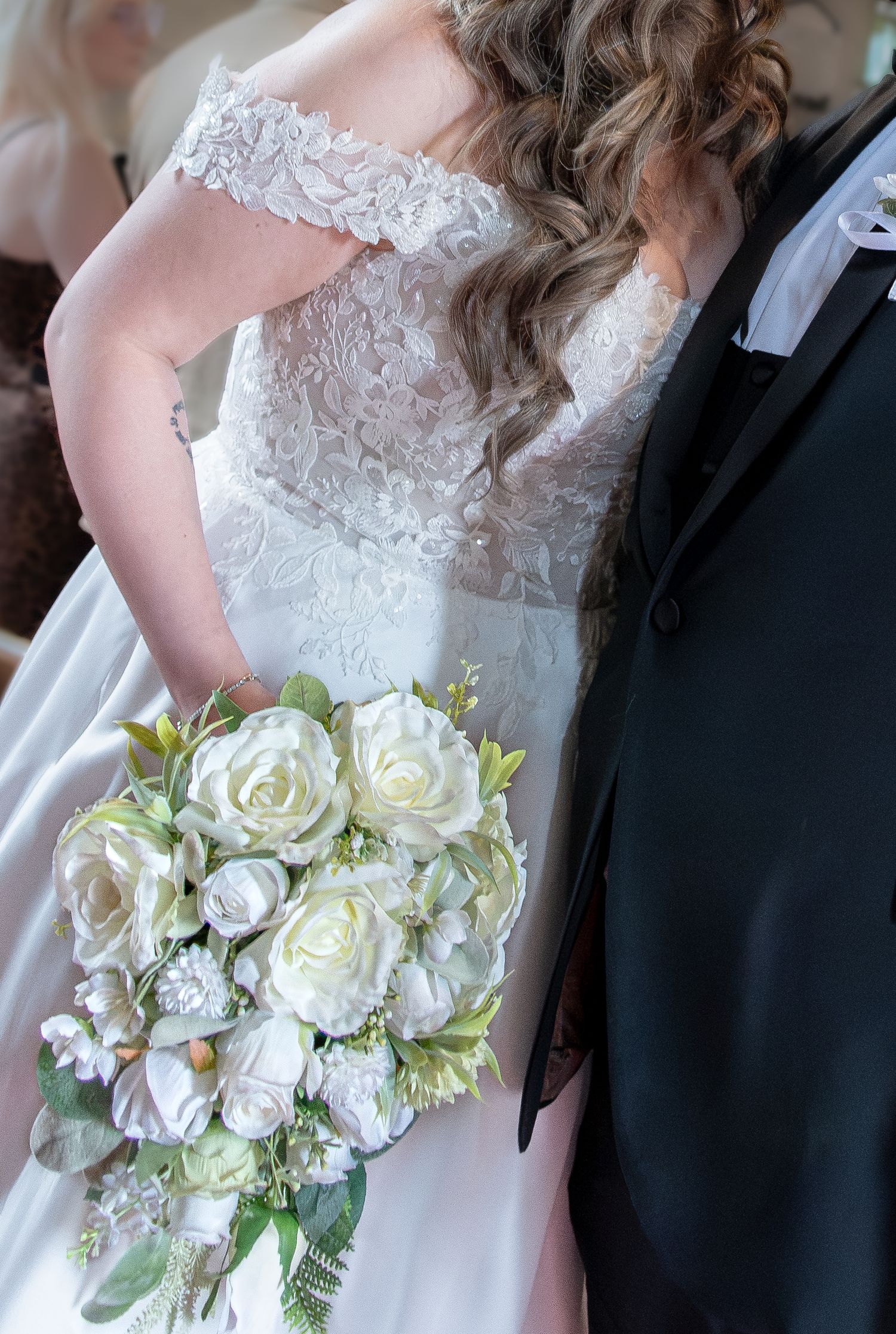 Bride in off-shoulder lace wedding gown holds white rose bouquet beside groom in black tuxedo.