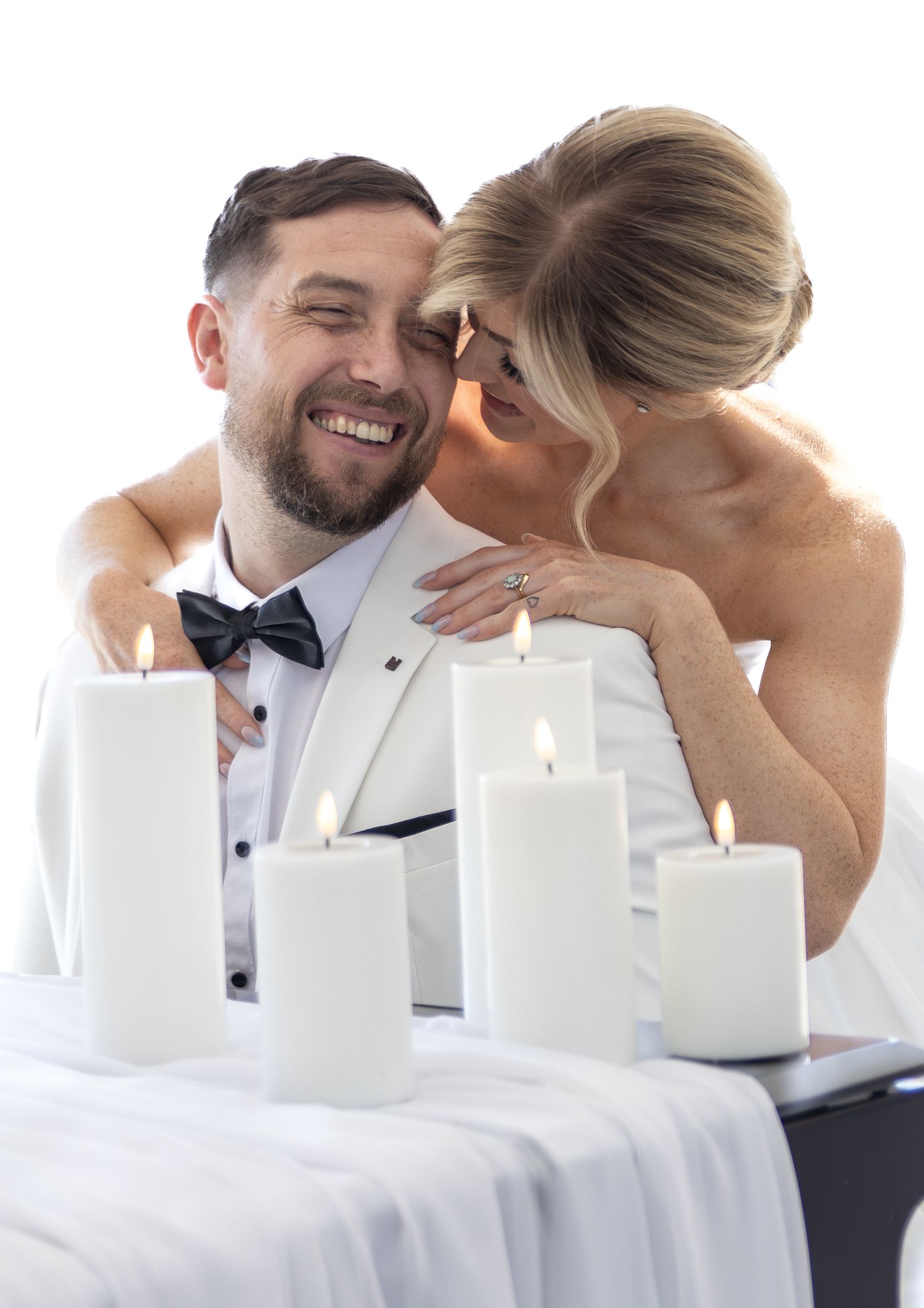 Happy groom in white tuxedo with bride whispering, surrounded by lit candles at wedding reception.