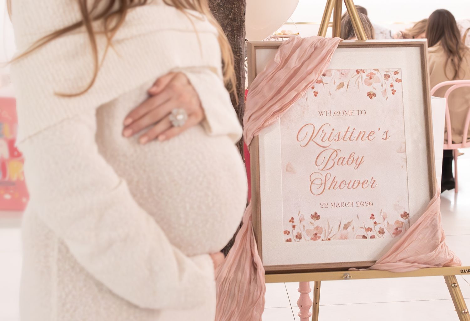 Pregnant woman in white outfit beside a floral pink baby shower welcome sign for Kristine's Baby Shower.