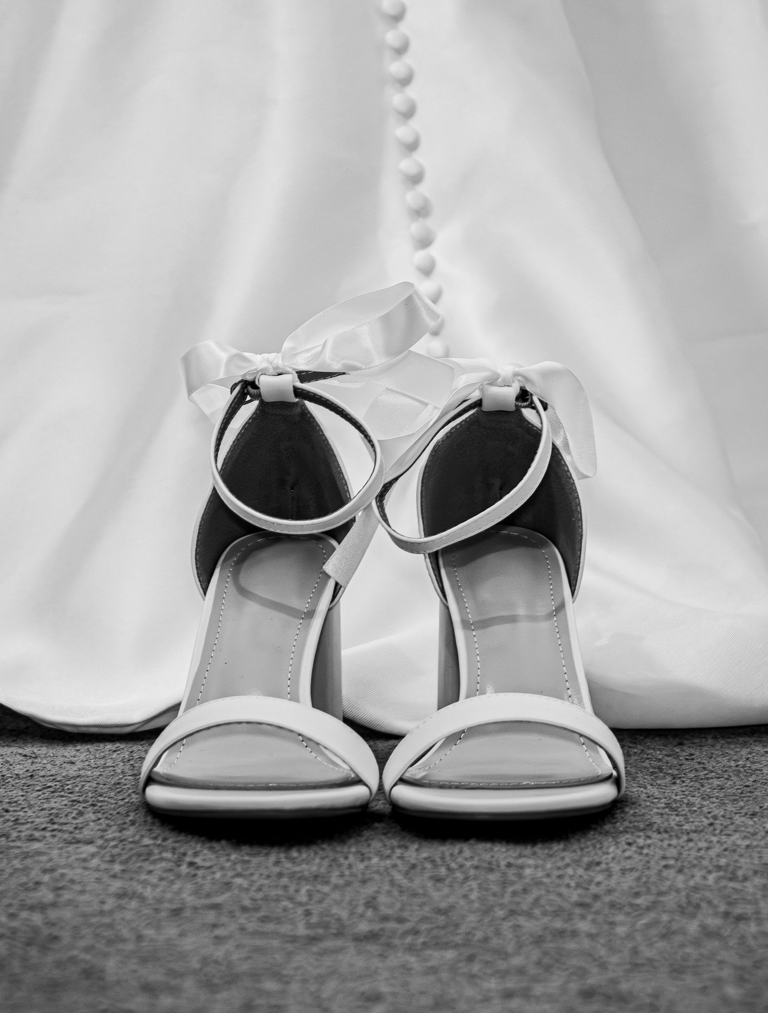 Black and white photo of elegant white strappy heeled sandals with bow detail placed in front of a wedding dress.