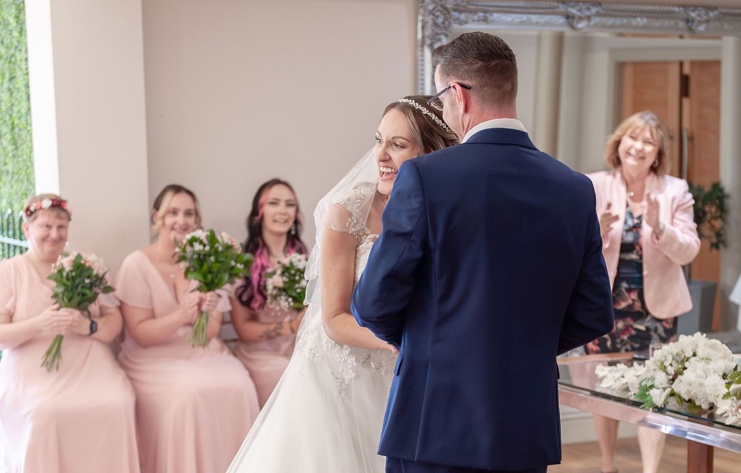 Bride and groom sharing a tender moment at the altar, surrounded by bridesmaids in pink dresses holding white bouquets.