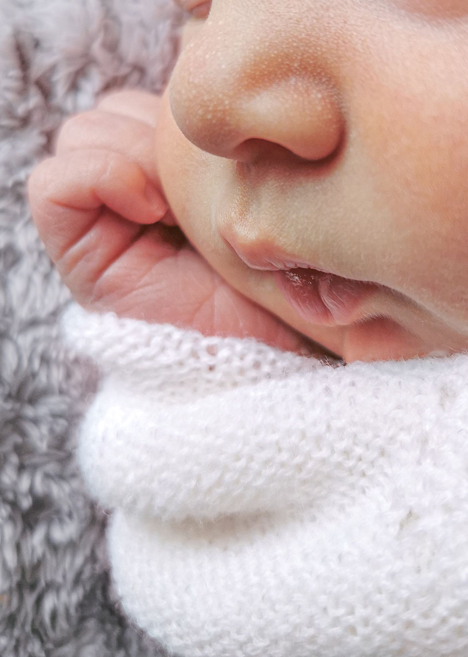 Close-up of a sleeping newborn baby's face and tiny hand wrapped in white knit fabric.