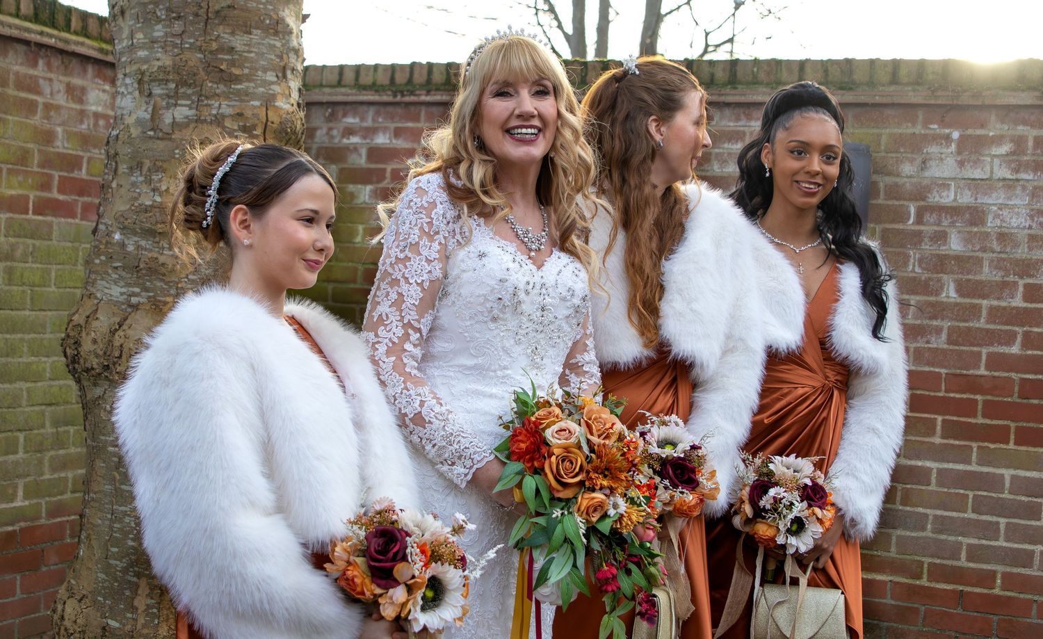 A group in white fur shawls holds autumn-colored bouquets against a brick wall backdrop at a fall wedding.