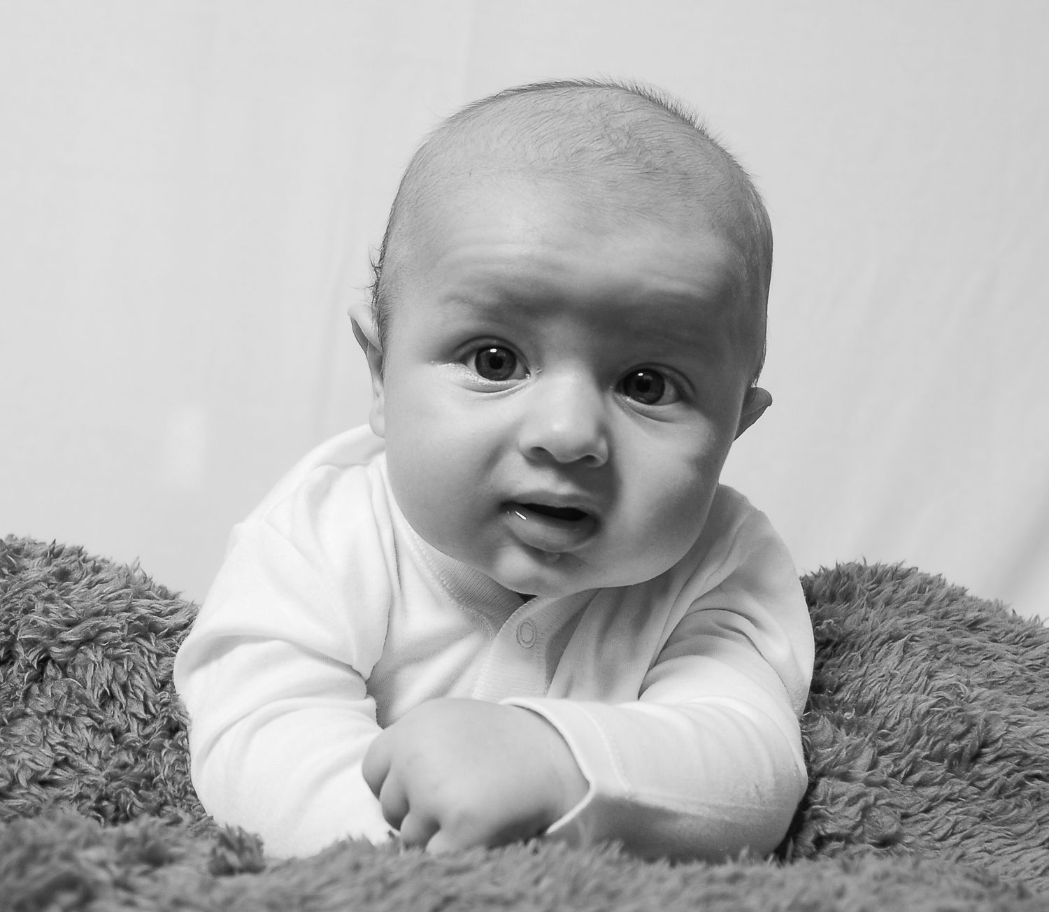 Black and white portrait of an adorable baby lying on a fluffy gray blanket, looking curiously at the camera.