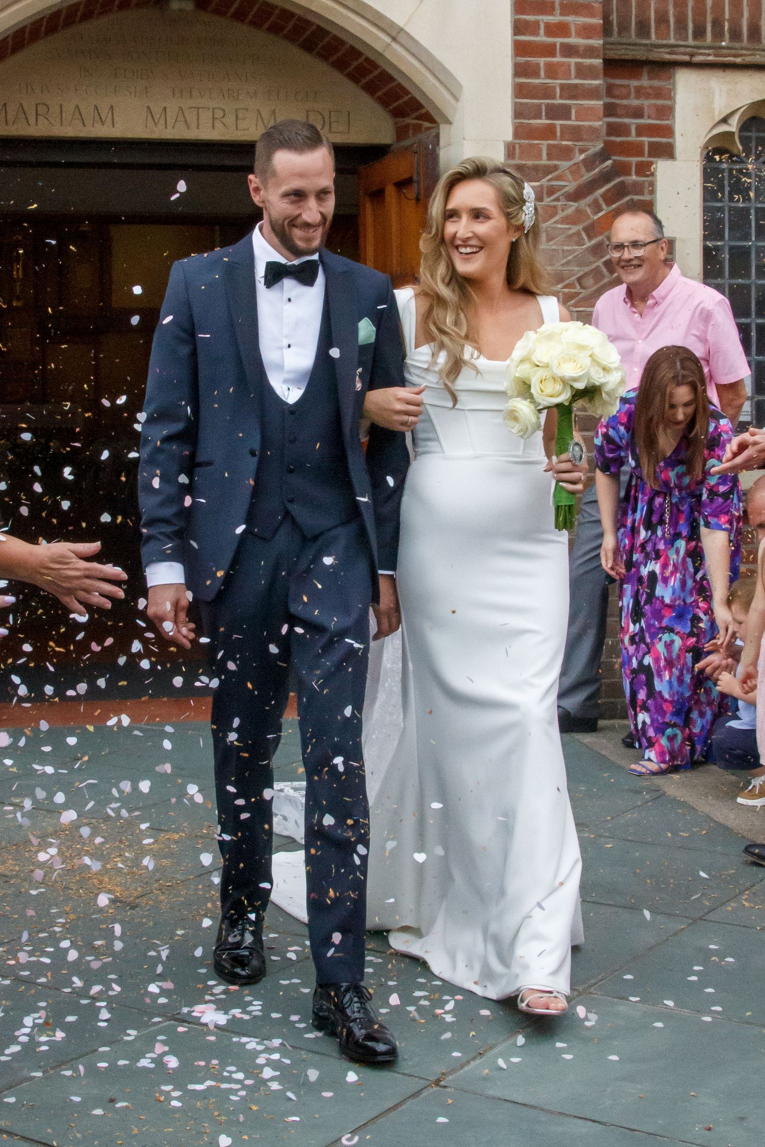 Wedding couple exits church under shower of confetti in elegant attire.