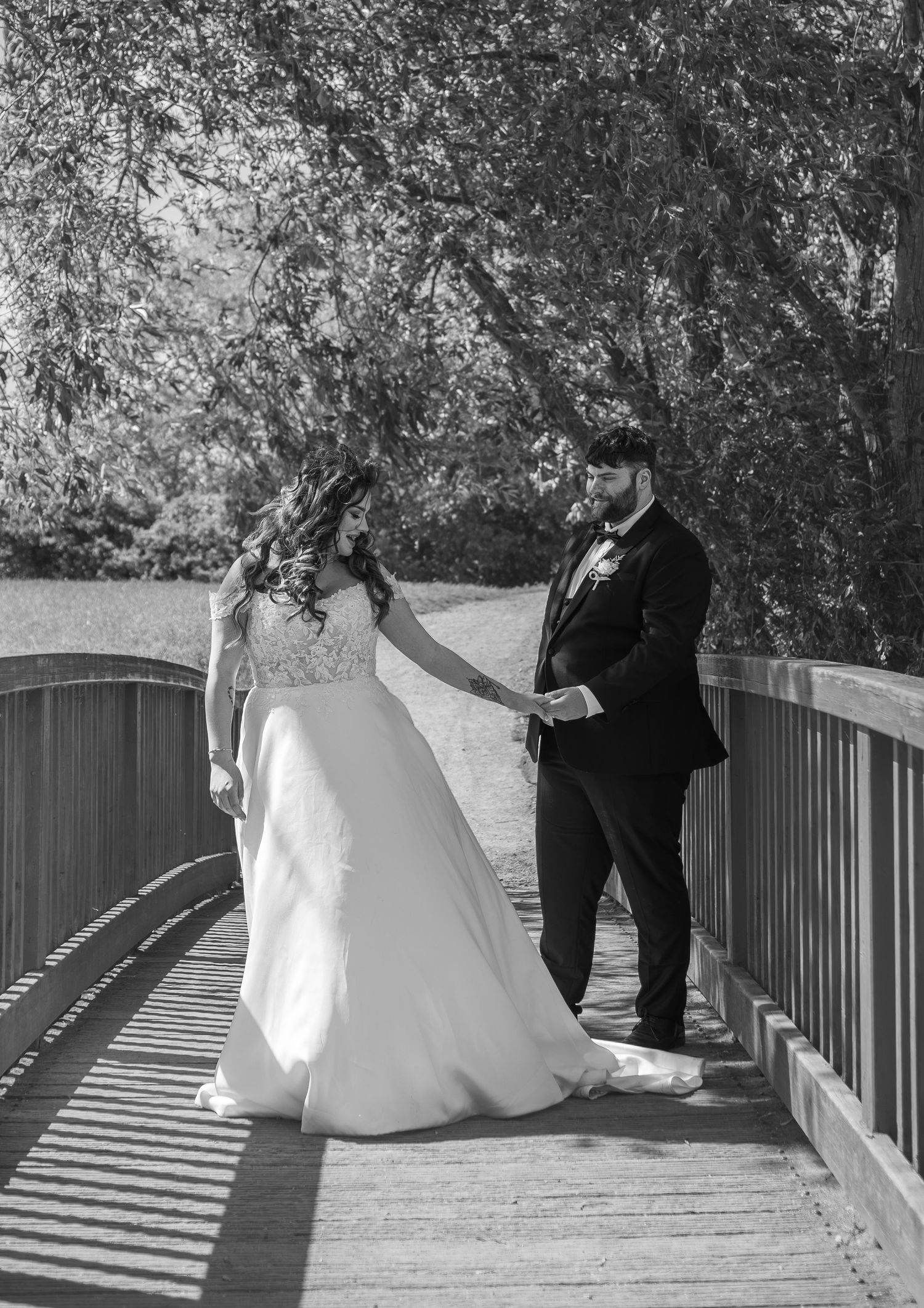 Black and white photo of bride and groom holding hands on a wooden bridge surrounded by trees on their wedding day.