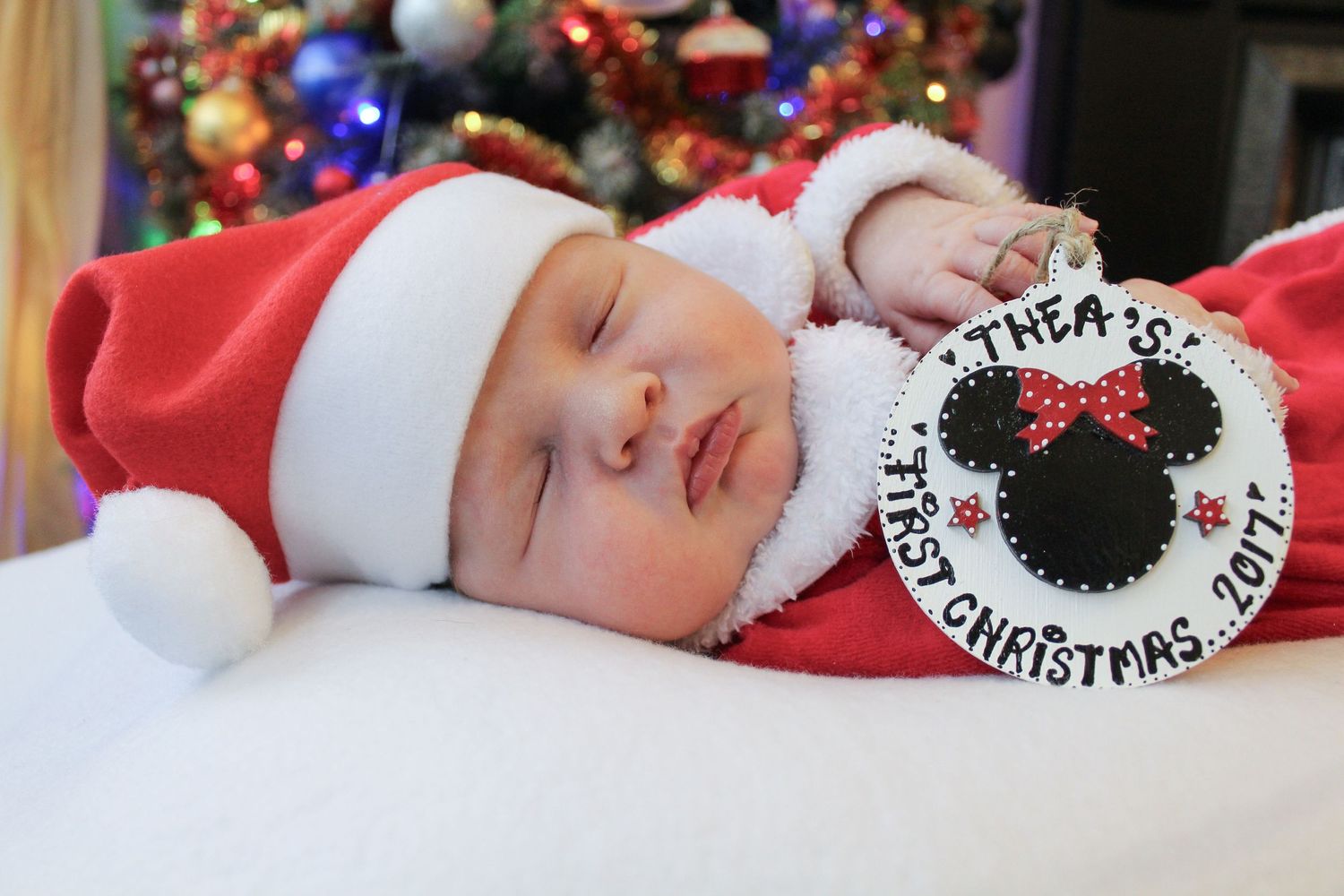 Newborn baby sleeping in Santa outfit with Minnie Mouse first Christmas ornament near decorated tree.