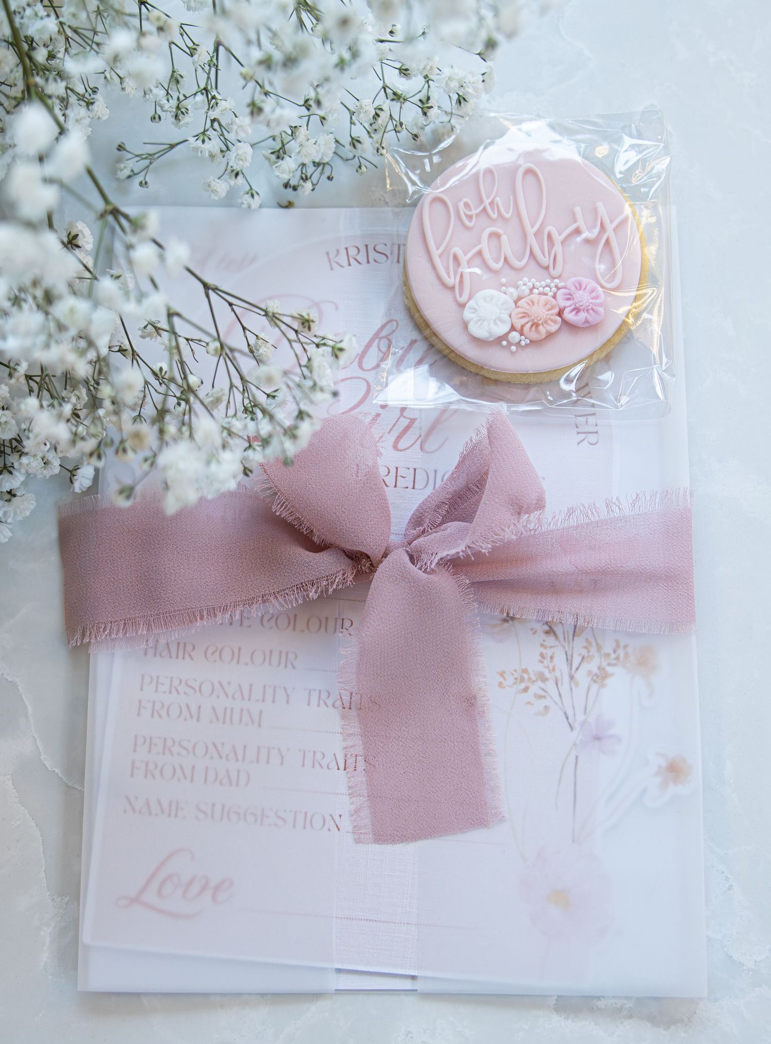 Pink baby shower gift set with decorated cookie, floral card, and dusty rose ribbon bow on white background.