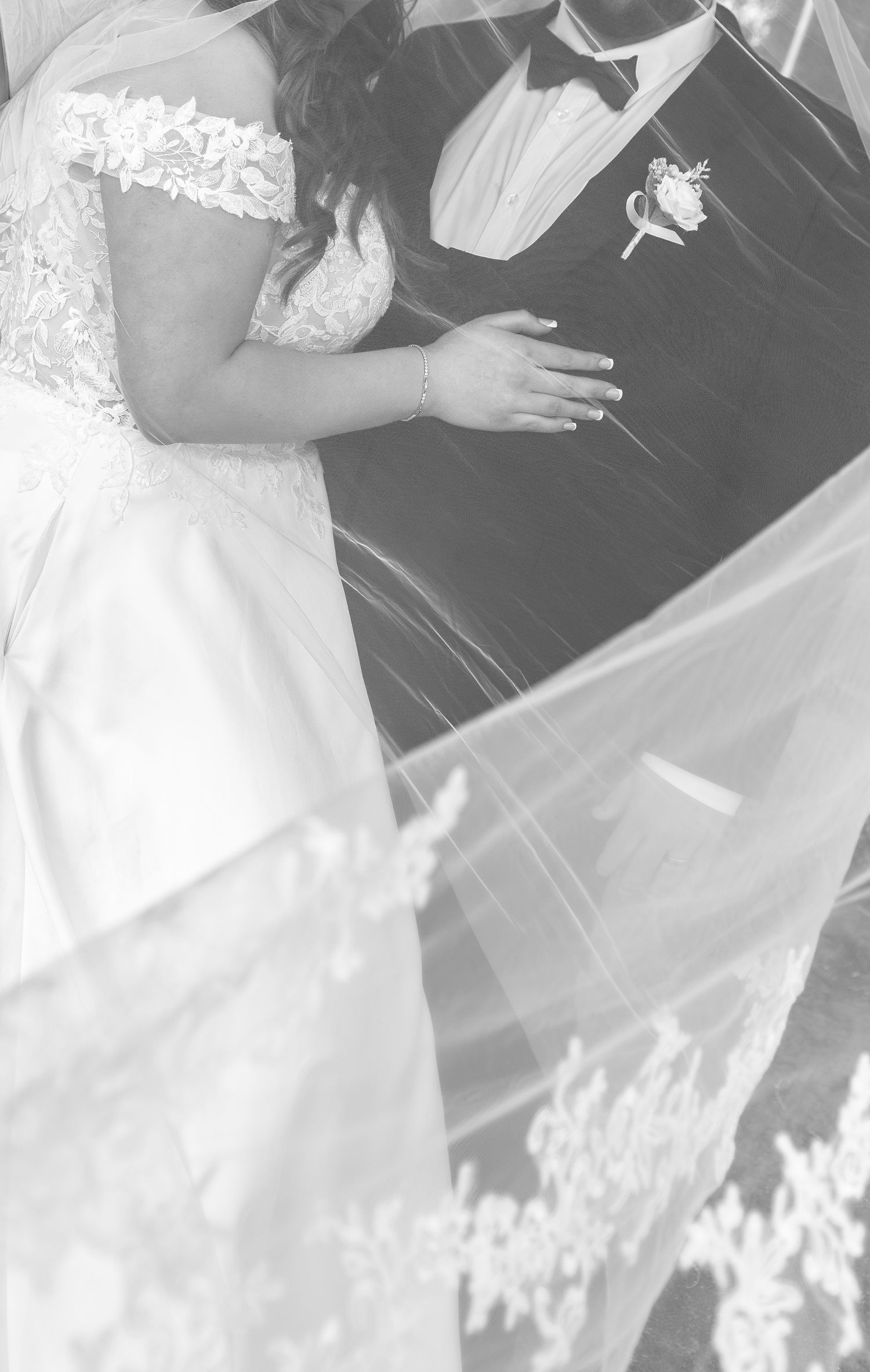 Black and white photo of bride in lace off-shoulder gown with flowing veil beside groom in tuxedo.