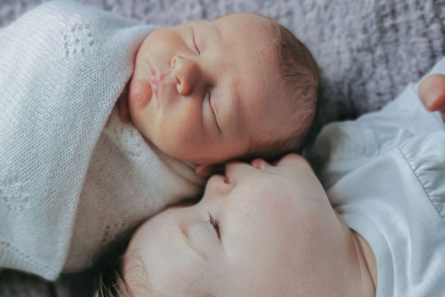 A newborn baby sleeping peacefully beside a young child, both resting on a soft gray blanket.