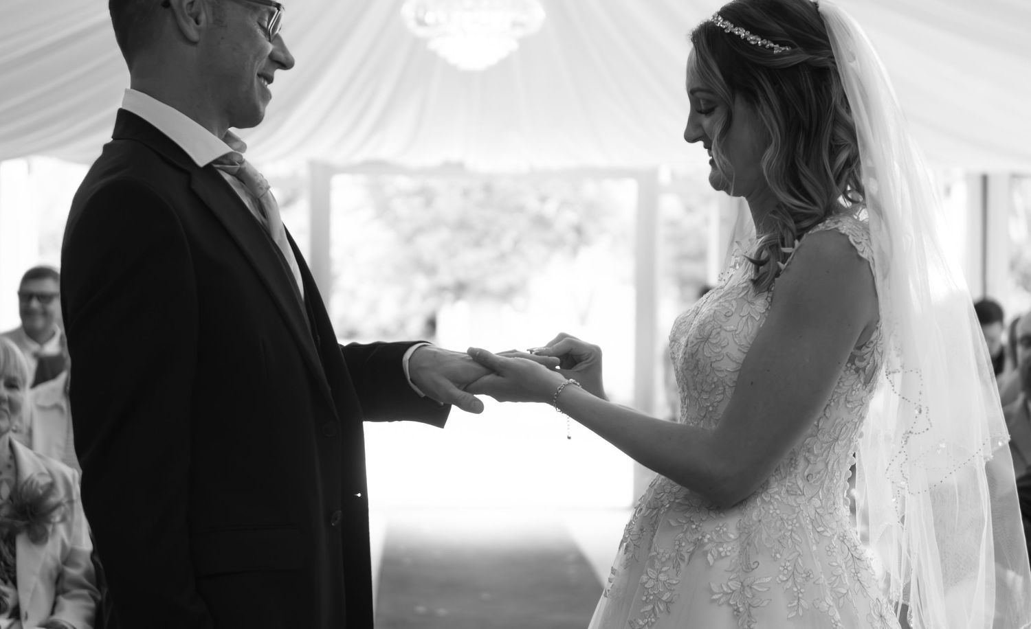 Black and white photo of a bride placing a wedding ring on the groom's finger during a ceremony.