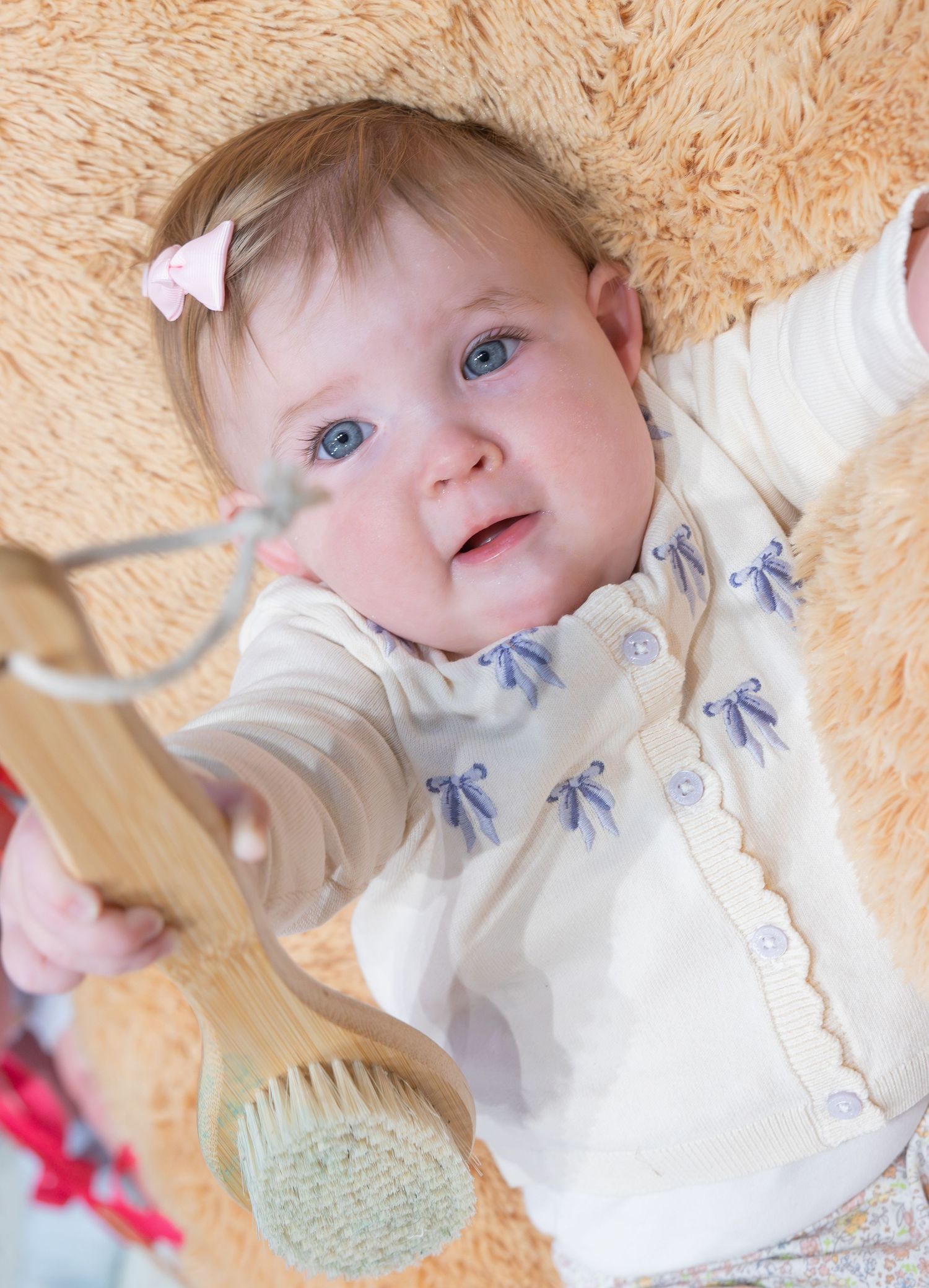 Blue-eyed baby girl in white cardigan with bow embroidery holds a wooden brush while lying on a fluffy beige carpet.