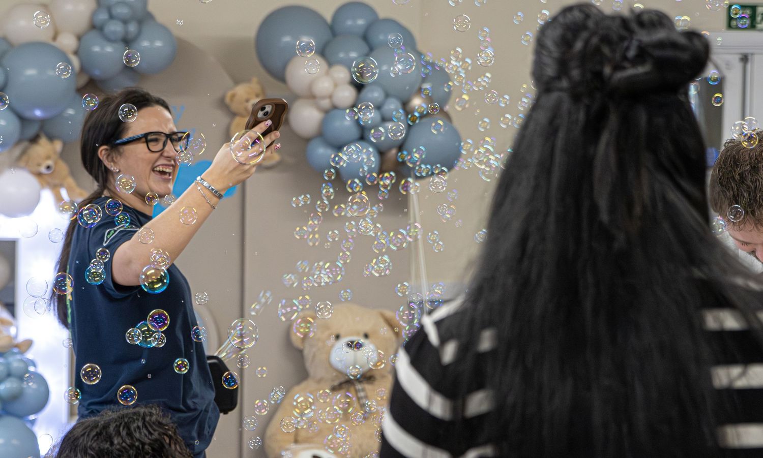 Woman blowing bubbles at a baby shower with blue balloons and teddy bear decorations in the background.