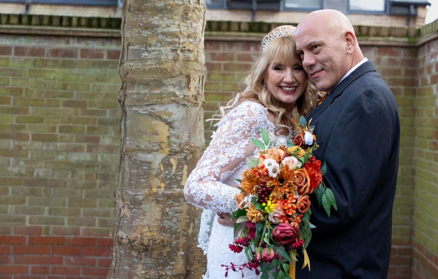 A couple shares a romantic moment in front of a brick wall while holding vibrant orange and red autumn bouquets.