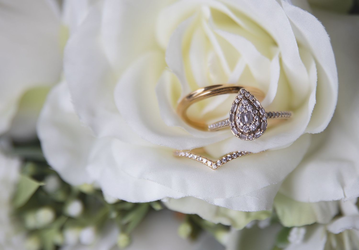 Elegant pear-shaped diamond halo engagement ring with gold band resting inside a white rose bloom.