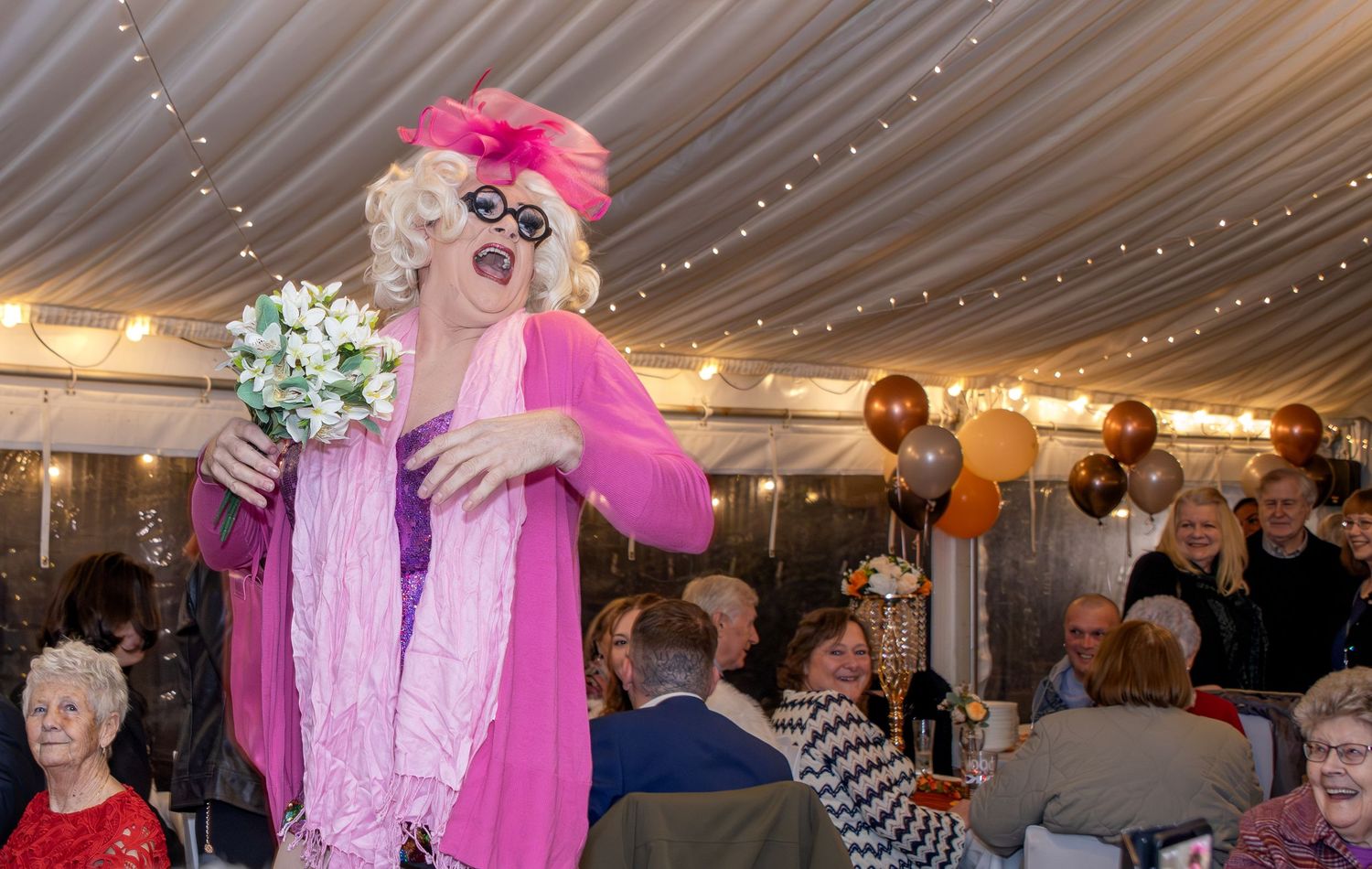 Drag queen in pink outfit holds bouquet at festive party with balloons and seated guests in marquee.