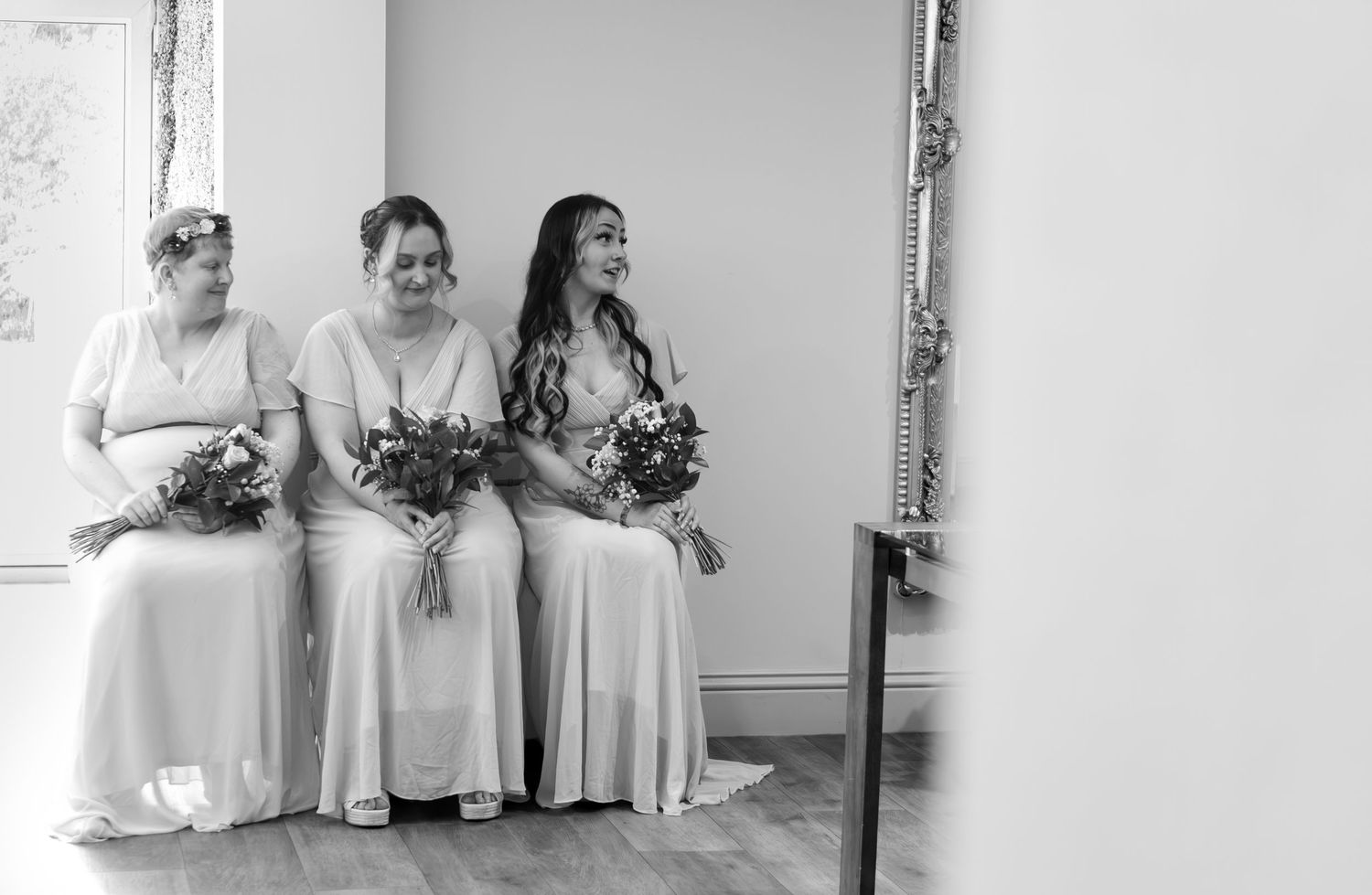 Three bridesmaids in white dresses holding floral bouquets seated together in a black and white wedding photo.