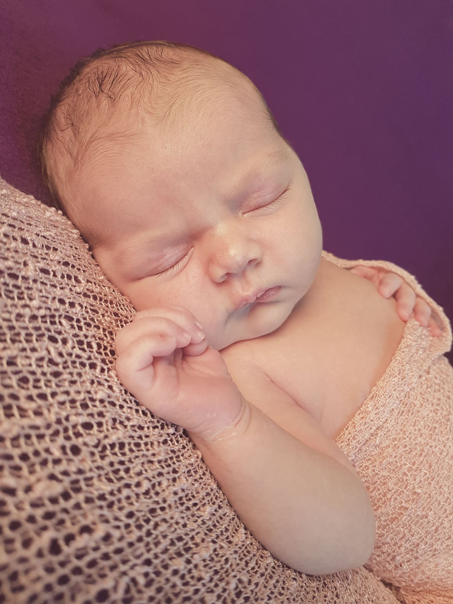 Peaceful newborn baby sleeping wrapped in a soft pink knit blanket against a purple background.