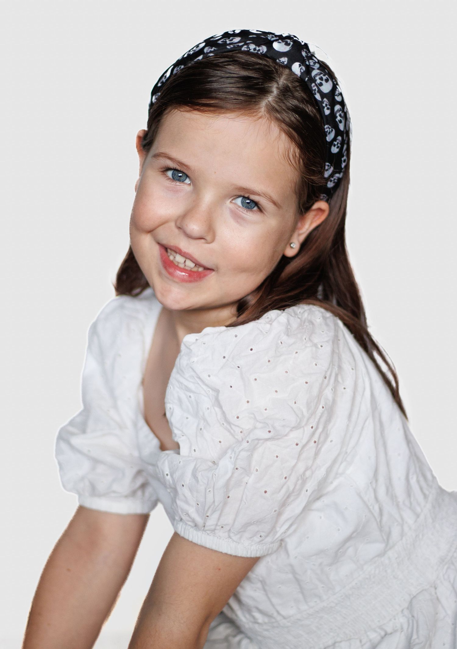 Young person in white eyelet dress with floral headband poses against white background.