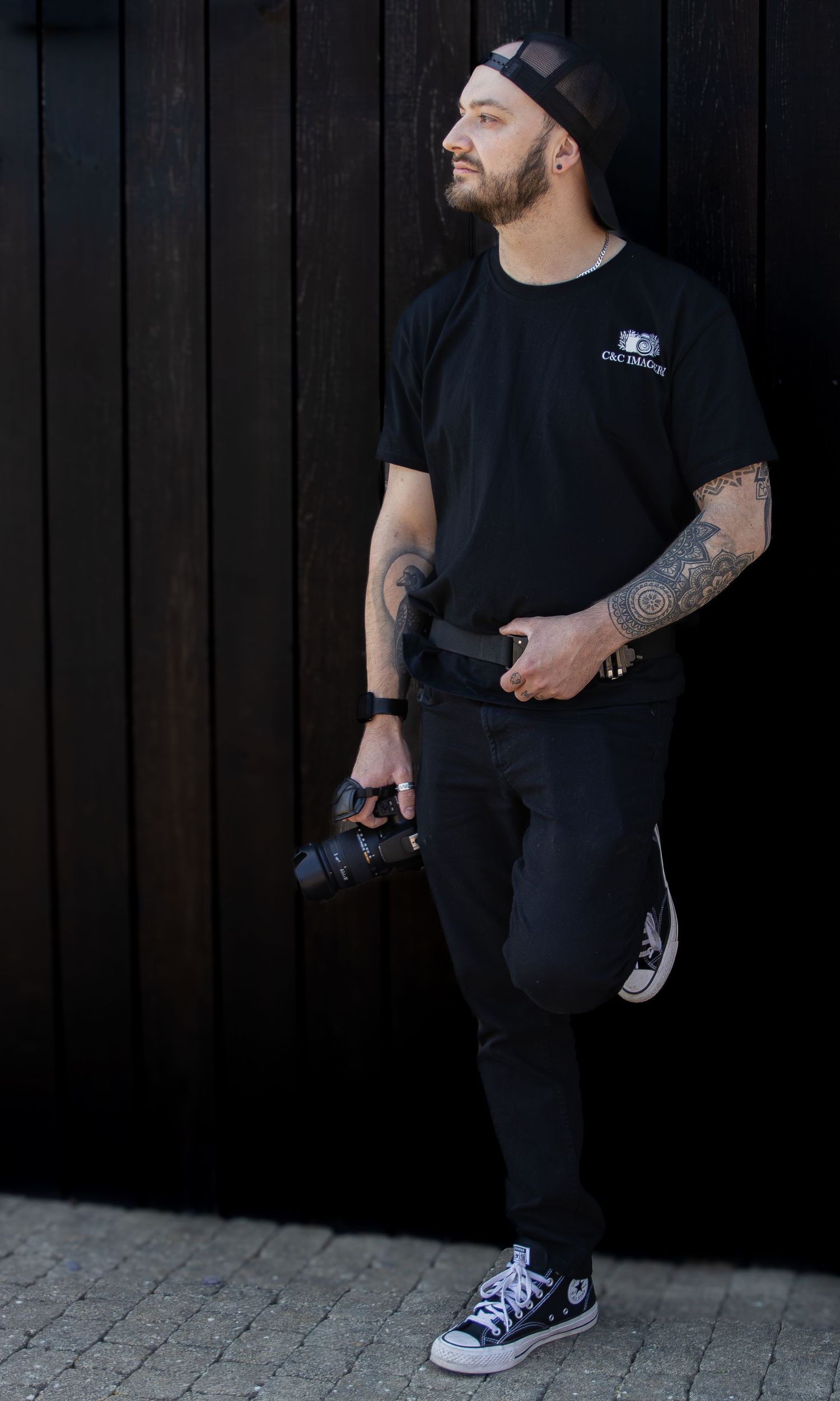 Tattooed male photographer in all-black outfit holding camera, wearing snapback cap and Converse sneakers against dark wood wall.