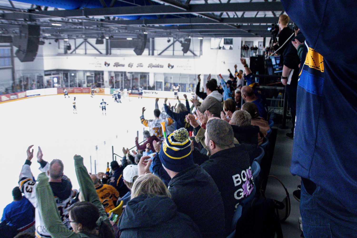 Spectators watch hockey game from elevated viewing area in indoor arena.