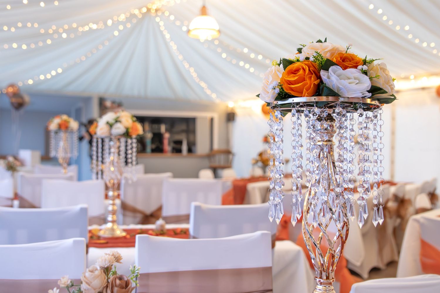 Elegant wedding reception decor with crystal chandelier centerpiece and white draping with string lights overhead.