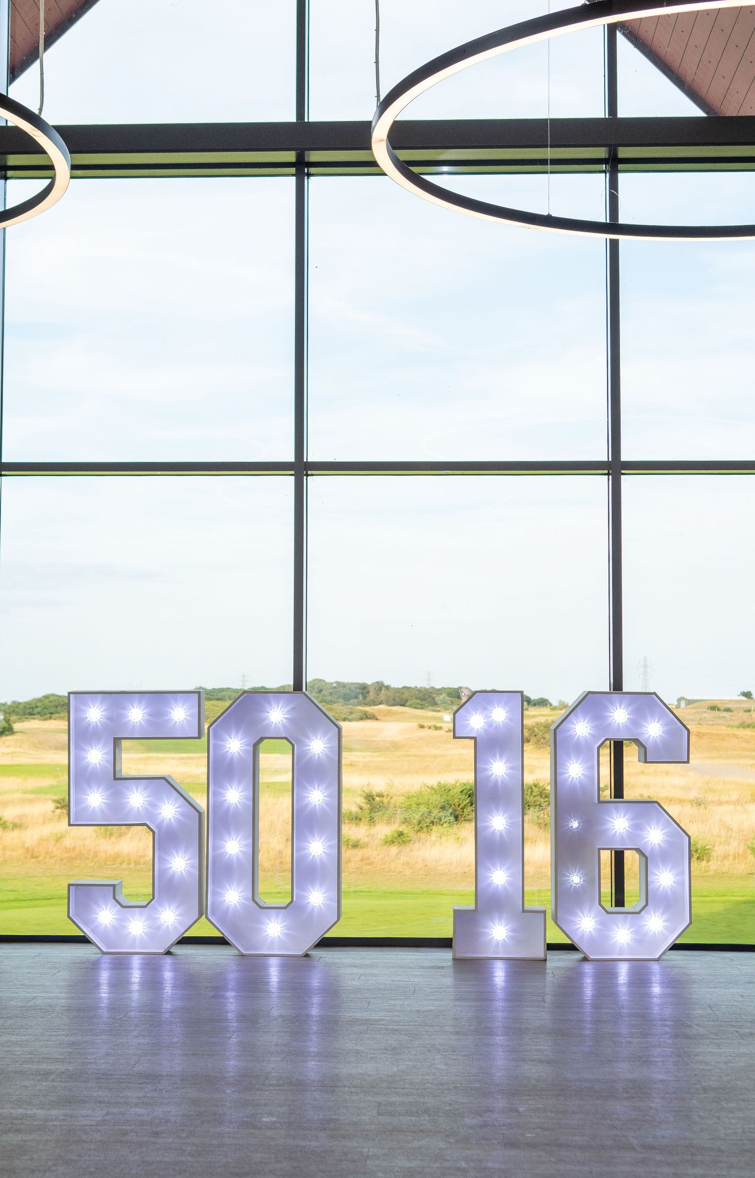 Illuminated marquee numbers 50 and 16 displayed in front of large windows overlooking a green landscape.