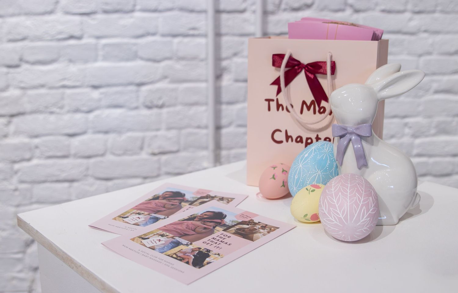 Easter-themed table display with ceramic bunny, pastel eggs, pink gift bag, and photo cards on white surface.