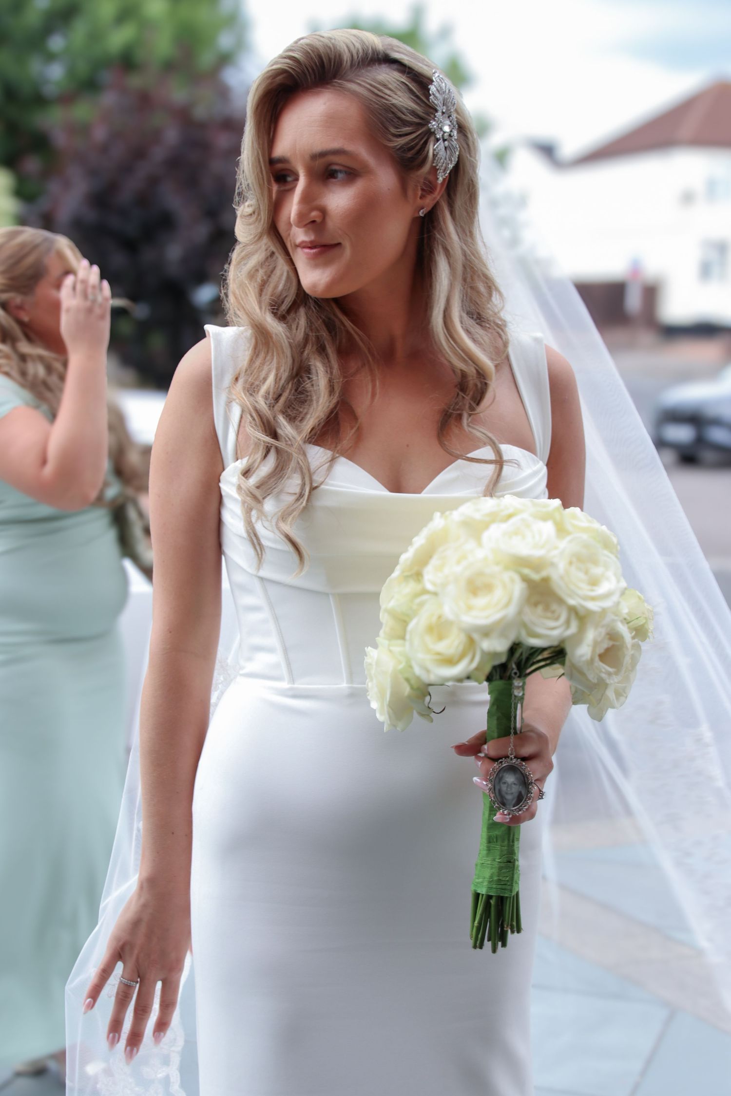 Bride in elegant white dress holds a classic bouquet of cream roses against an outdoor backdrop.