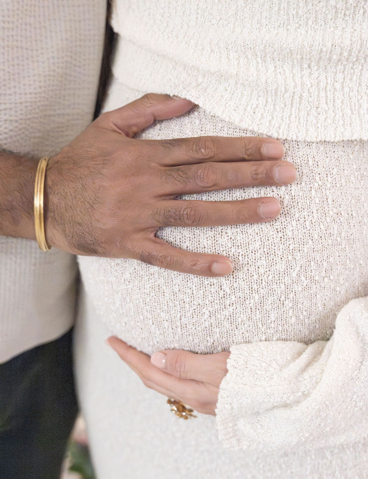 Close-up of hands on a pregnant belly, one wearing a gold bracelet, the other with a floral ring, both in white.