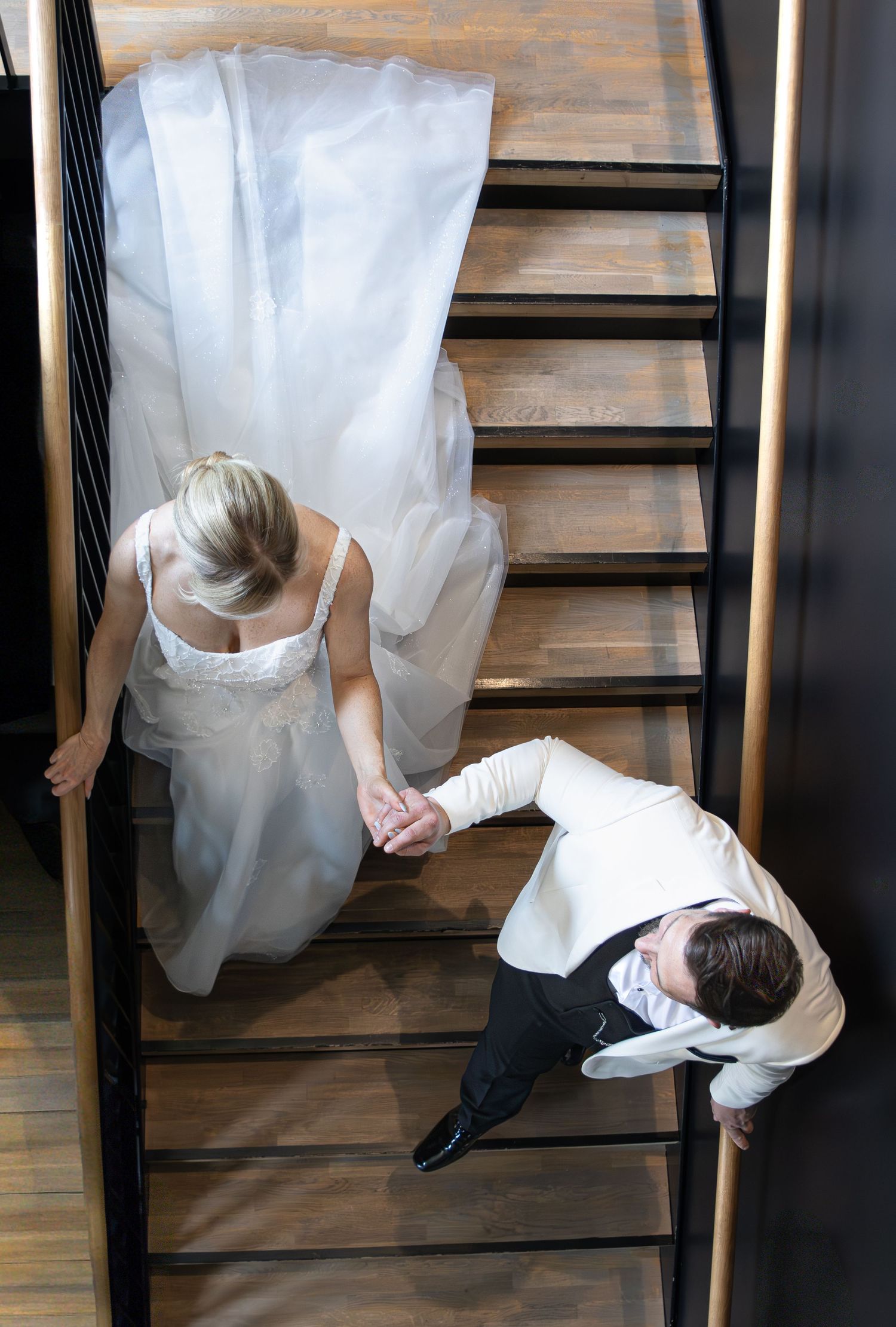 Aerial view of bride in white gown and groom in tuxedo holding hands while descending wooden staircase.