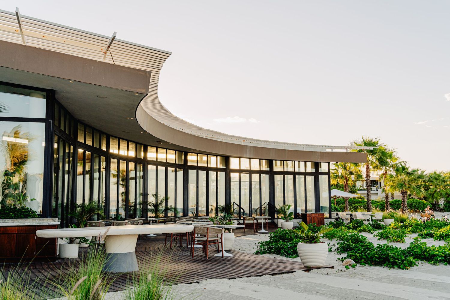 A modern architecture building with curved glass walls and outdoor walkway surrounded by landscaping and palm trees.