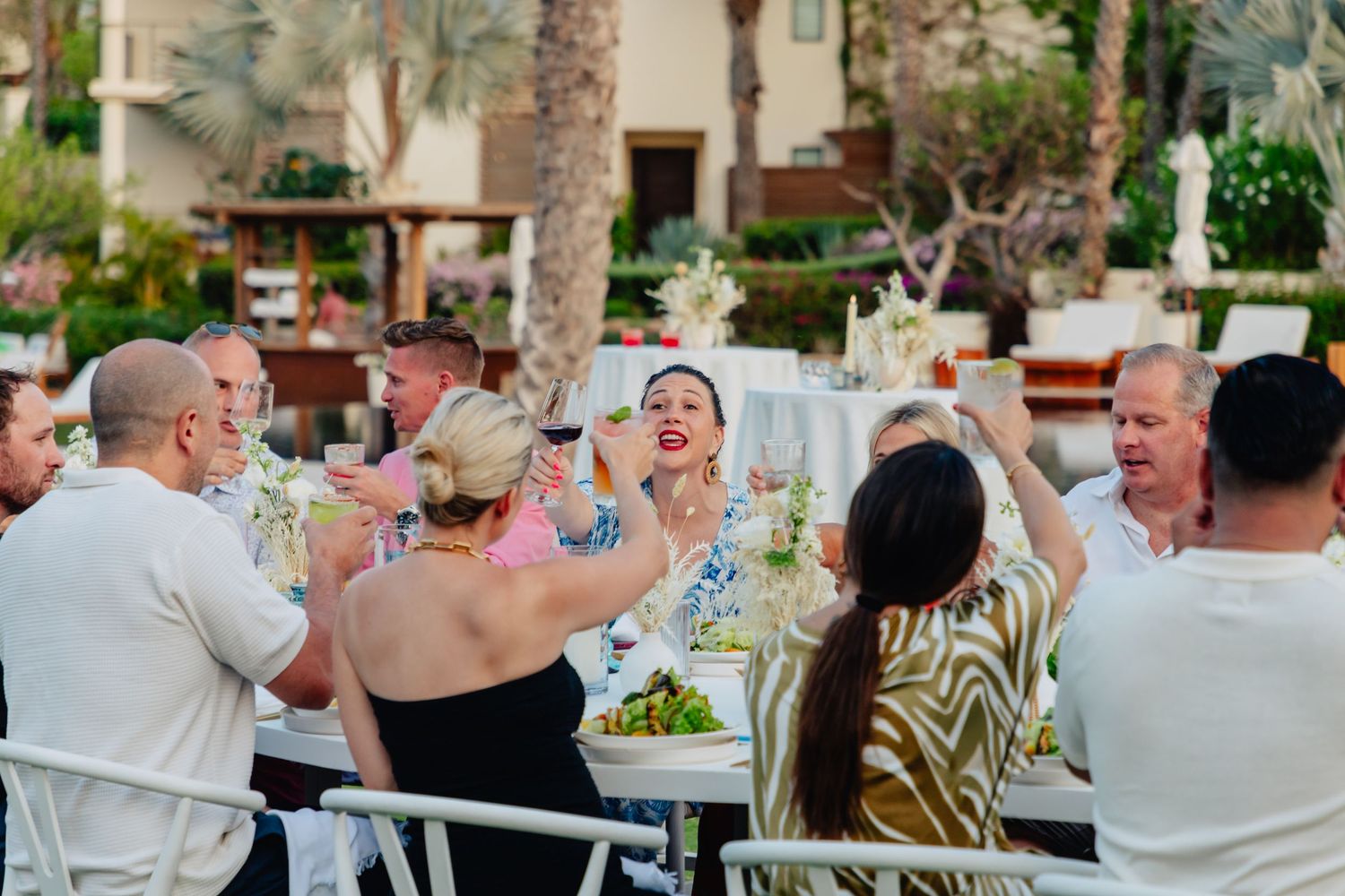 Wedding guests raise their glasses in celebration during an outdoor reception at a garden venue.
