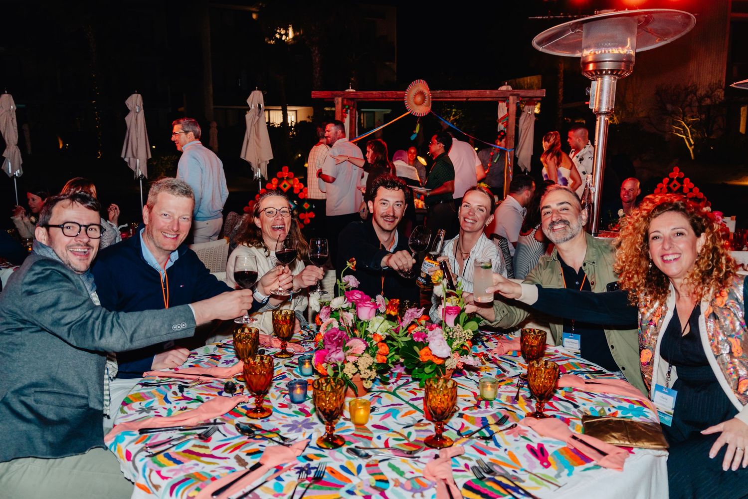 People gather around decorated tables at a festive dinner party with colorful table settings and flowers.