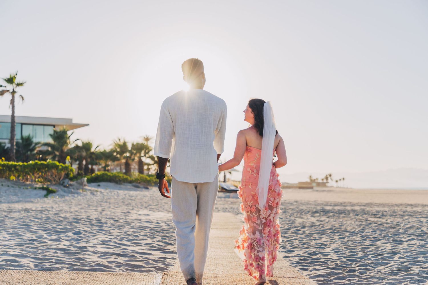 A romantic couple walks along a pristine beach at sunset with palm trees and ocean waves in the background.