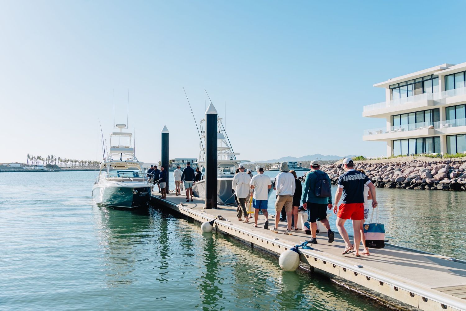 People walking along a wooden pier on a sunny day with boats and buildings visible in the background.