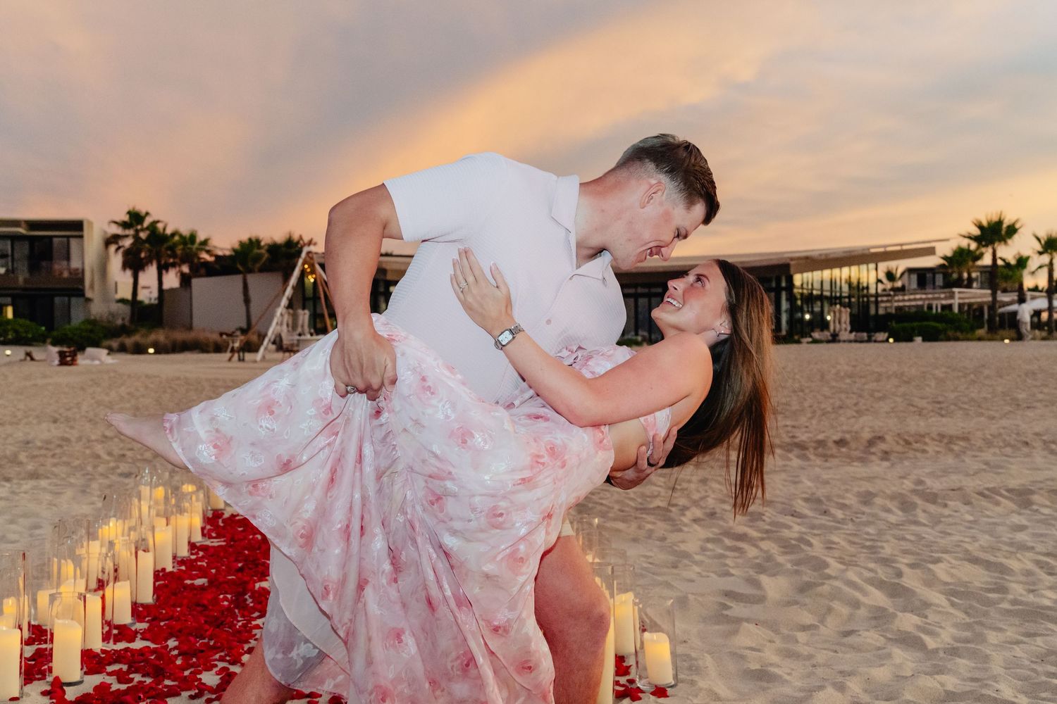 Romantic beach wedding ceremony setup with candle lanterns lining a rose petal aisle at sunset.