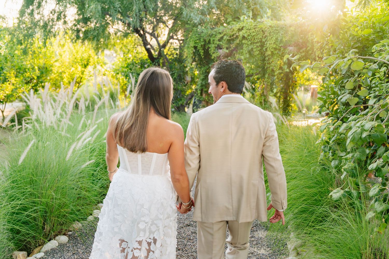 A couple in wedding attire walks through a lush garden path during golden hour sunset.