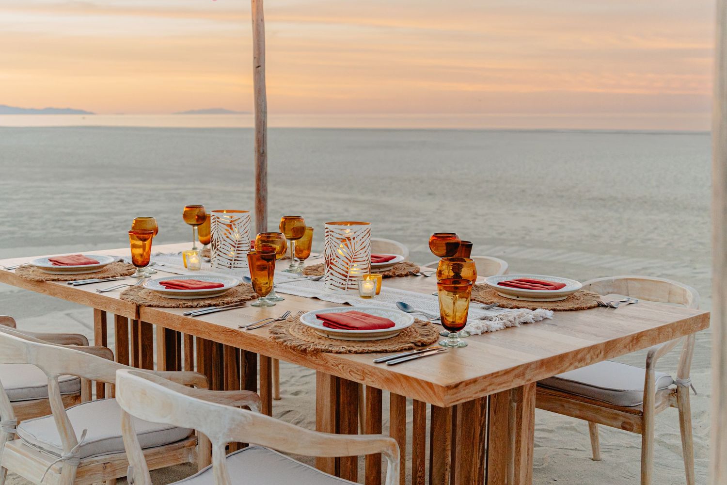 Wooden dining table set for dinner with amber glassware against a pastel sunset seascape.