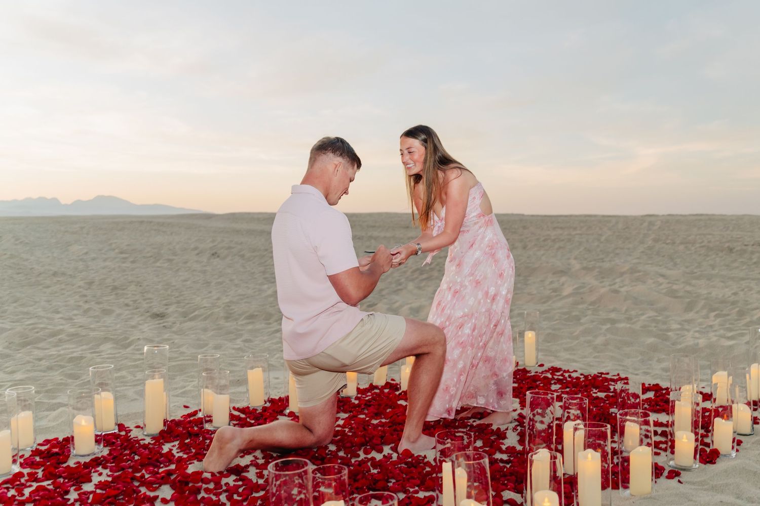 A romantic beach proposal setup with candles and rose petals at sunset.