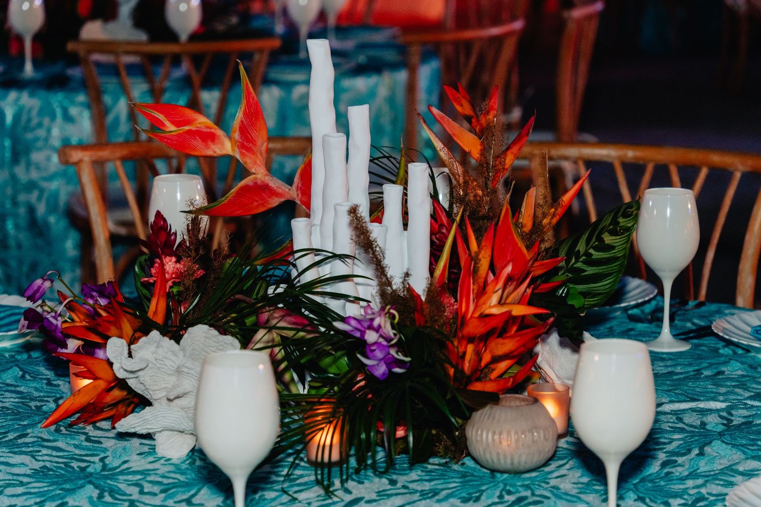 Tropical floral centerpiece with red birds of paradise and white candles on turquoise tablecloth.